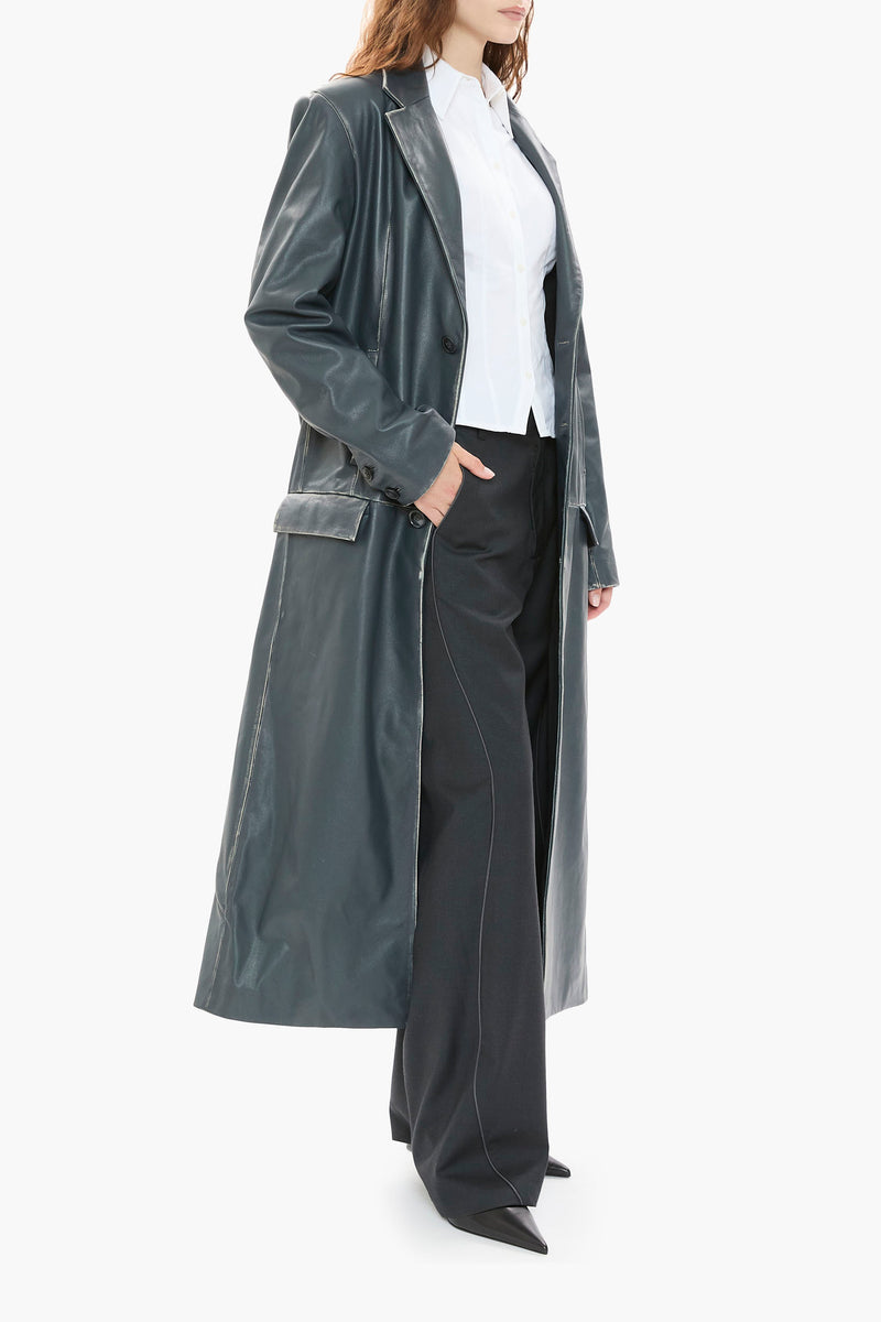 Person wearing a long dark blue leather coat over a white shirt and black pants on a white background